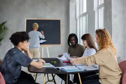 Group of students studying together at a table while one writes on a blackboard in a bright classroom.